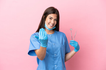 Dentist woman holding tools isolated on pink background inviting to come with hand. Happy that you...