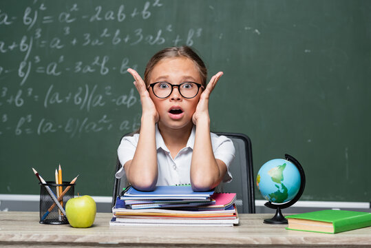 shocked schoolkid with open mouth looking at camera near notebooks and blurred chalkboard
