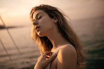 Portrait of tender young lady with blonde fluffy hair, closed eyes in white clothes and round earrings against sunlit sea and sky background during beautiful pastel pink sunset