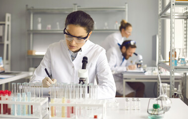 Portrait of young serious female scientist making notes sitting at lab desk with chemical...