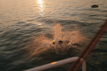 Man jumping into sunlit golden-blue sea at sunset during summer vacation and water splashing beautifully, shimmering in sun
