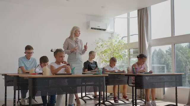 In Elementary School Class: Enthusiastic Teacher walks between Rows of Bright Diverse Children, Explains Lesson. Group of Smart Multiethnic Kids Learning New Stuff. Side View Tracking Shot