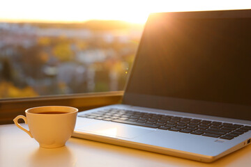 Hot coffee cup and laptop on table against the window at sunrise. Cozy workplace in home office, concept of remote work and earning online