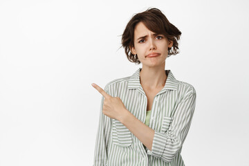 Disappointed frowning girl, sulking upset, pointing finger at upper left corner and furrow eyebrows skeptical, standing against white background