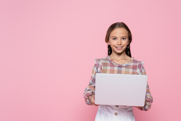 preteen girl smiling at camera while holding laptop isolated on pink