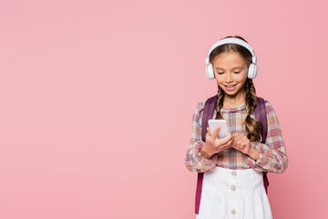 Smiling schoolchild in headphones using cellphone isolated on pink