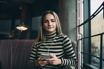 Portrait of Caucaisan millennial blogger in electronic headphones looking at camera, young hipster girl with modern cellphone gadget in hands posing while listening audio book in cafe interior