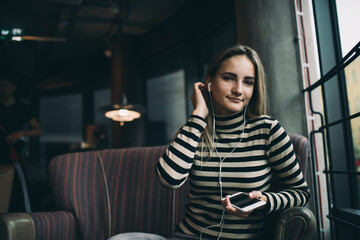 Fototapeta na wymiar Portrait of Caucaisan millennial blogger in electronic earphones looking at camera, beautiful hipster girl with modern mobile gadget in hand posing while listening audio book in cafe interior