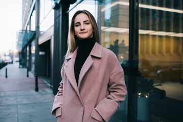 Half length portrait of attractive female model dressed in stylish pink coat standing at urban setting in city and looking at camera, Caucasian fashionista posing at street during spring daytime
