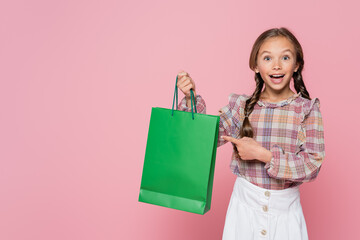 amazed and happy child pointing at green shopping bag isolated on pink