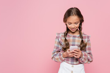 smiling girl in checkered blouse messaging on smartphone isolated on pink