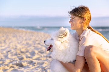 A teenage girl hugs a dog, a white Samoyed husky, and looks into the distance at the sea coast. Pets, friendship with animals concept.