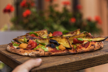 Woman's hand holds a wooden tray with pinsa hummus and glazed eggplantst