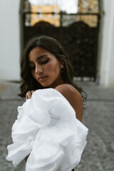 Tender brunette with long curly hair, smooth tanned body in white off-shoulder top, vintage earrings, sensitively posing on street and closing eyes 