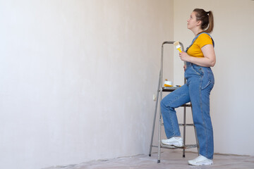 A pensive woman with brush and ladder looks dreamily in a room with an unpainted wall with copy space. Independent single female makes DIY repairs in her apartment. 