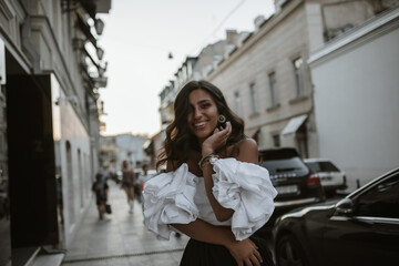 Joyful lady with brunette curly hair and tanned body, in retro outfit of white off-shoulder blouse, dark pants, stylish earrings, walking on street, smiling and looking into camera