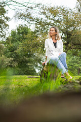Woman posing sitting on a wooden log in a green area of ​​Asturias, Spain.The girl is wearing jeans, a white jacket and transparent shoes.Photo with blur in the foreground and shot in portrait format.