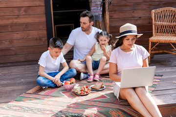 Freelance mom works with a laptop sitting on the porch during a family vacation, in the background...