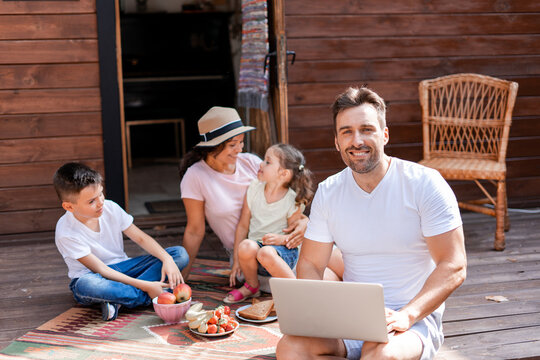Smiling man works during family vacations, uses WiFi and 5G uses a laptop, digitalization of his work processes and the opportunity to work sitting on the porch of a wooden house