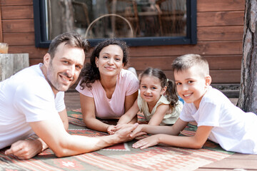 A loving, friendly and caring family lie together near a wooden house and hold hands. Mom, dad, daughter and son spend time together during summer vacation.