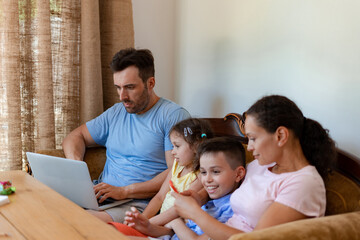 Family of four, all sitting on a comfortable sofa and using their electronic gadgets, the daughter watching the father work during the family weekend, the mother playing games on the phone