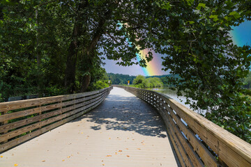 a long winding boardwalk along the river with a wooden rail along the sides with vast still river water, lush green trees with blue sky and clouds at Roswell Riverwalk Boardwalk in Roswell Georgia