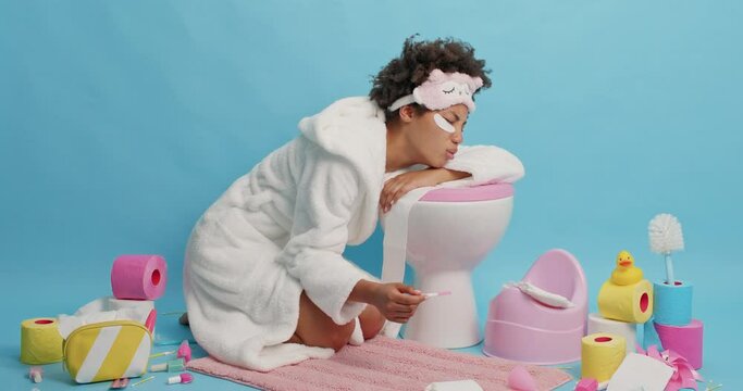 Unhappy curly haired woman leans on toliet bowl dissatisfied with pregnancy result wears white bathrobe and sleepmask has stressed expression isolayed over blue background. Poor contraception