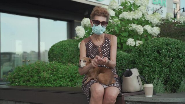Ederly woman in medical mask sits on bench in city and disinfects her dog dachshund hands and paws with antiseptic. Senior female masked with pet use anitizer in park during quarantine covid 19.