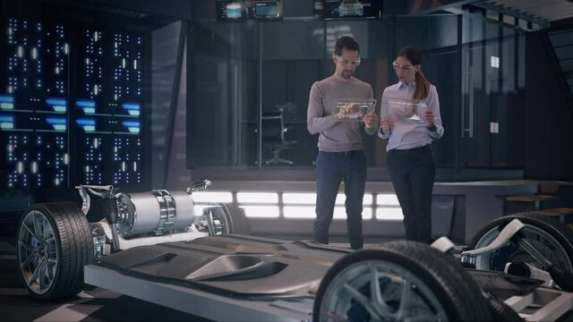 Mockup of Electric Vehicle: Automotive Professional Engineers working on design of Electric Car using Futuristic Transparent Touchscreen Tablets inside High-tech facility. Electric car chassis.