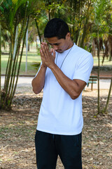 Young Brazilian man in a public park, with his hands close to his face, in a salute position.