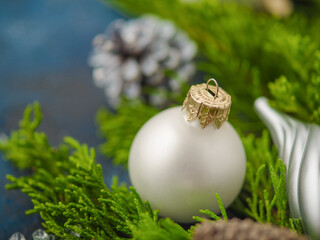 In the foreground is a glass white Christmas ball against a background of green coniferous branches. In the background there are bumps. Blue background. Christmas composition on a blue background.