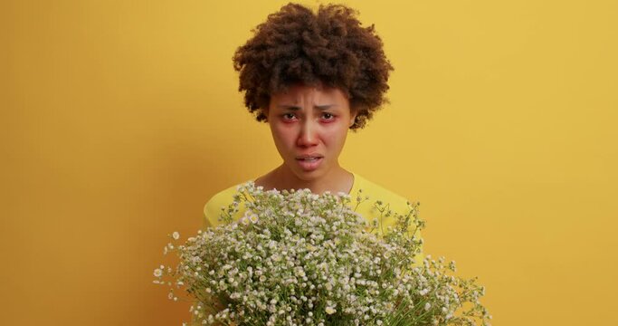 Frustrated curly haired young woman sneezes constantly suffers from allergic reaction on pollen holds bouquet of wildflowers feels unwell has respiratory symptoms poses against yellow background
