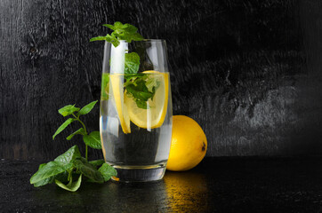 A large glass of water with lemon, mint leaves. A glass of lemonade on a black textured background.