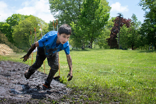 Young Boy Covered In Mud Running Through Fun Muddy Outdoor Obstacle Course In A Field.