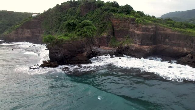 Aerial view of waves break on rocks in a blue ocean. Sea waves on beautiful beach 4k shot.  ocean waves crashing against an empty stone rock cliff from above. El Zonte beach, El Salvador