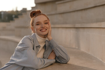 Frontal portrait of joyful young ginger chilling at sunny city. Adorable lady with collected hair and light clothes, smiling, looking into camera and posing outdoors