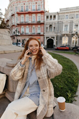 Fototapeta premium Urban portrait of young busy lady in city. Cute ginger with long smooth hairstyle, shirt, light plaid coat, talking on phone while sitting against buildings background