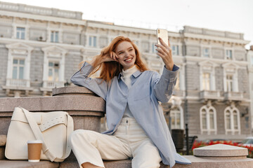 Fototapeta premium Charming young lady making selfie in city against buildings background. Attractive foxy girl with blue shirt, white pants and stylish accessories, holding phone and posing