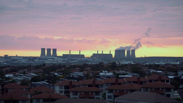 A beautiful early morning sunrise (or sunset) of orange and pink clouds over a coal-fired power station and blocks of houses in a suburban setting. Wide shot.