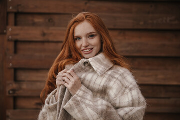 Beauty portrait of young foxy lady against wood background. Charming long-haired girl with freckles, nude makeup, plaid coat, smiling, holding collar and posing outdoors