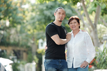 Portrait of Asian mature woman smiling at camera while standing outdoors together with her elder son