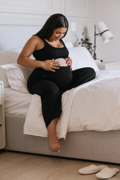 Pregnant Carribbean Indigenous woman practicing self care and putting lotion on baby bump in bed