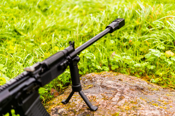 Rifle standing on a bipod on a rock in the grass after a rain. Close up view of a stone level, focus on the flame arrestor