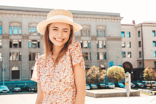 Young beautiful smiling hipster woman in trendy summer sundress. Sexy carefree woman posing on the street background in hat at sunset. Positive model outdoors. Cheerful and happy.Winking
