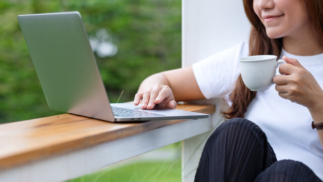 Closeup image of a beautiful young asian woman drinking coffee while using and working on laptop computer in the outdoors