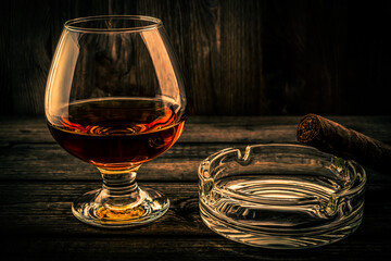 Glass of brandy and glass ashtray and cuban cigar on a old wooden table. Focus on the cuban cigar