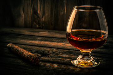 Glass of brandy and cuban cigar on an old wooden table. Angle view, focus on the glass of brandy