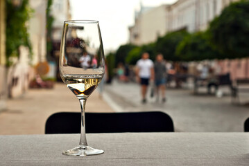 Glass of white wine close up. Alcoholic drink on a wooden table on a background of the city. Transparent wine on the summer terrace of the restaurant. Blurred people in the background.