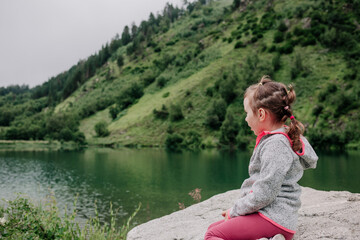 a little girl in a gray pullover sits on a rock and looks at the lake