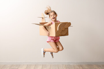Little girl with cardboard airplane jumping near light wall
